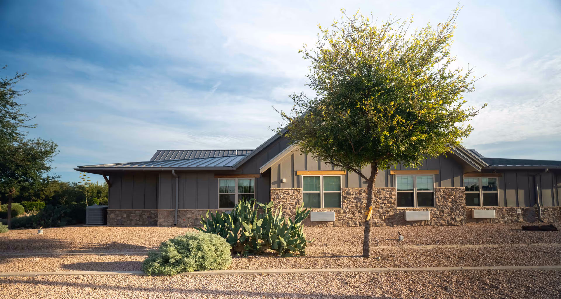 Exterior view of a single-story building with stone and gray siding, surrounded by a gravel landscape with a tree and shrubs under a partly cloudy sky.