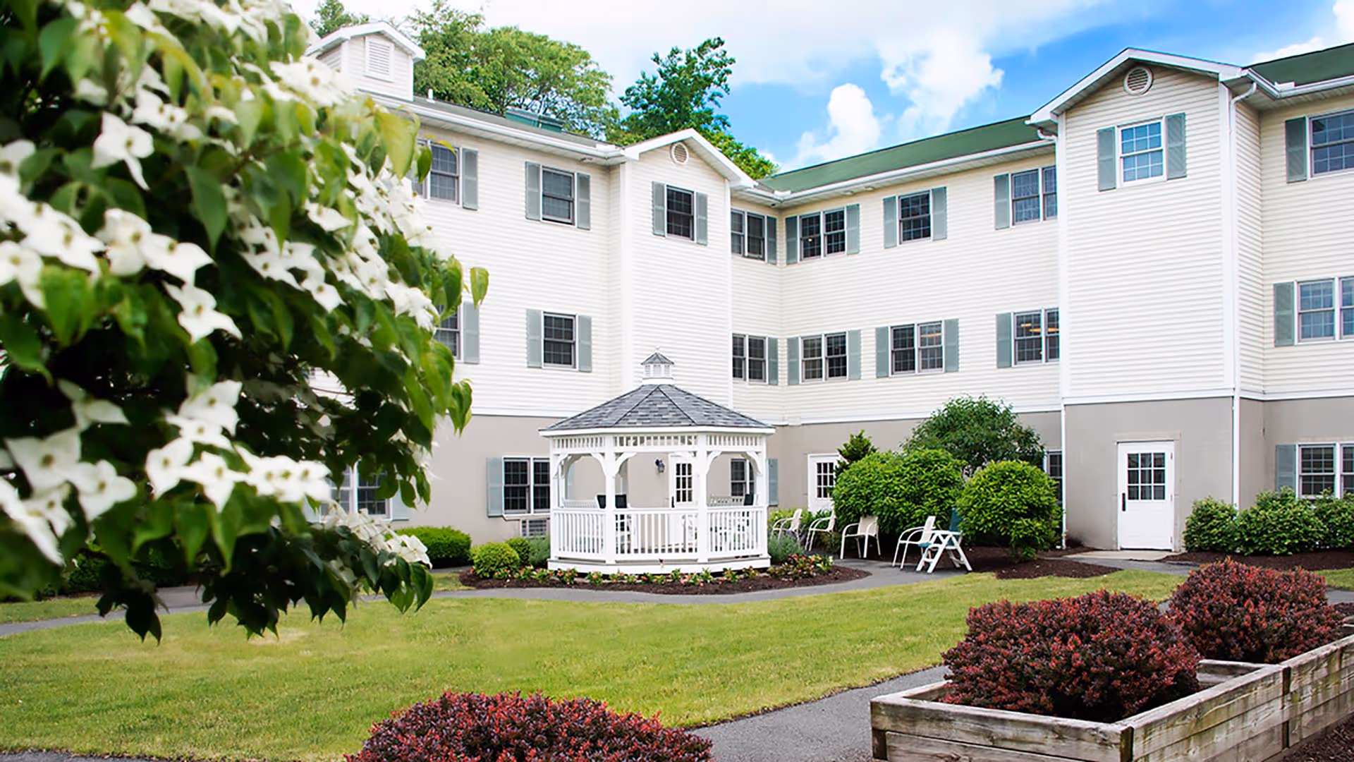 Outdoor courtyard area of a senior living facility featuring a white gazebo in the center surrounded by green grass, bushes, and flowering plants. The three-story building with white siding and multiple windows encloses the courtyard. Several chairs are placed near the bushes along the building walls under a partly cloudy blue sky.