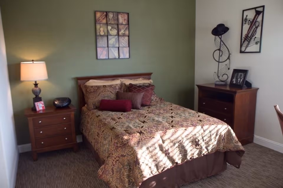 A cozy bedroom with a double bed covered in a patterned bedspread and multiple pillows. There is a wooden nightstand with a lamp, a decorative bowl, and a small framed photo on the left side of the bed. On the right side, there is a wooden dresser with a framed photo and a decorative music-themed sculpture with a hat on top. The walls are painted green and white, and there is a framed artwork above the bed.