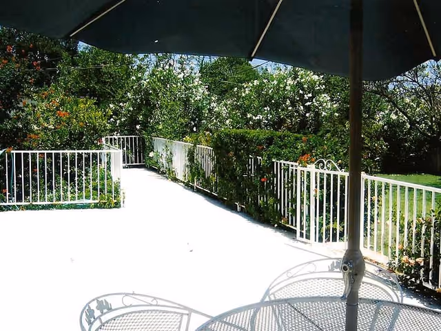 Outdoor patio area with white metal railing and green bushes along the perimeter. There is a large umbrella providing shade over a white metal table and chairs. Trees and blue sky are visible in the background.