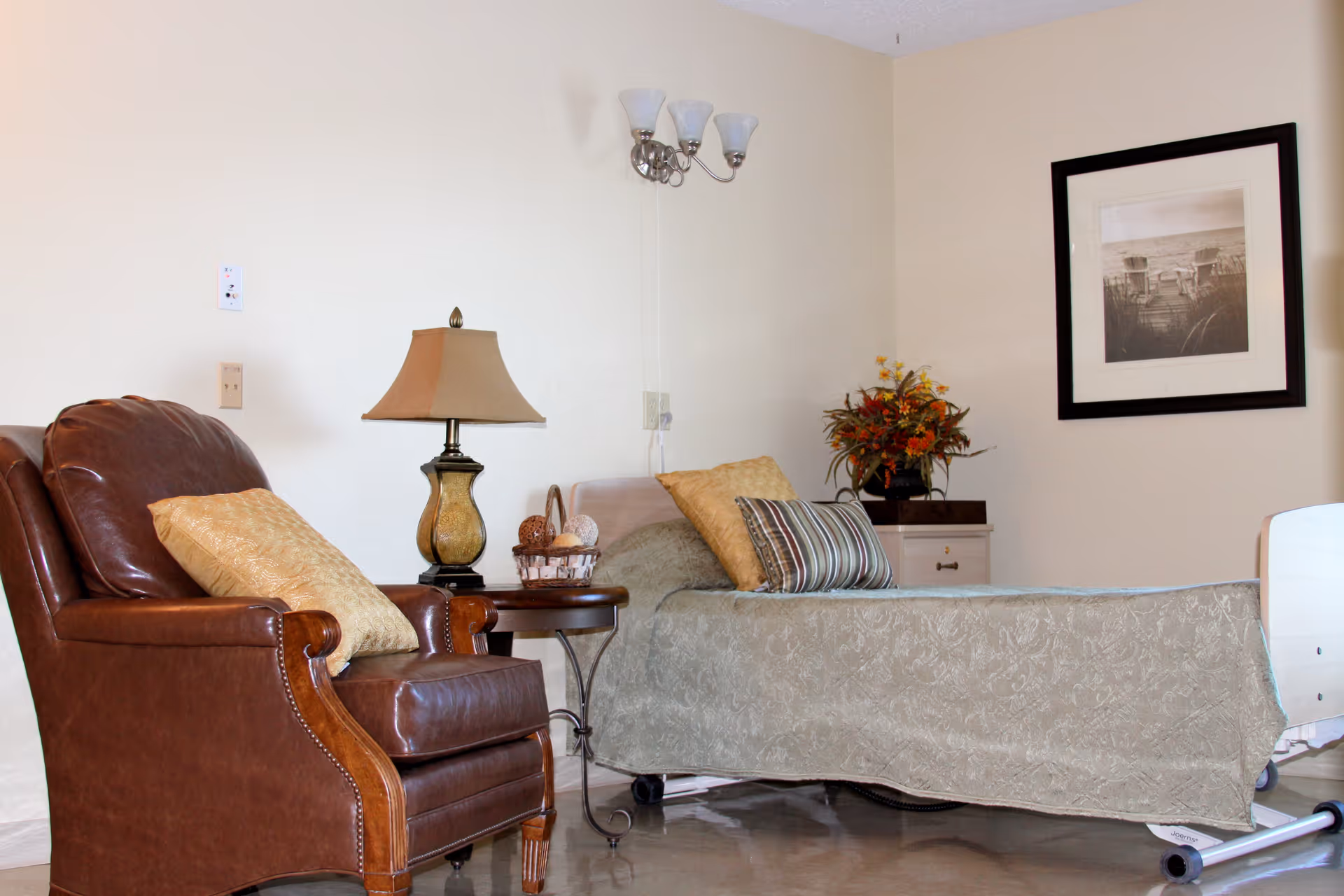 A cozy nursing center room with a brown leather armchair with a yellow pillow, a side table with a lamp and decorative items, and a hospital bed with patterned bedding and multiple pillows. There is a framed black and white picture on the wall and a floral arrangement on a small cabinet.