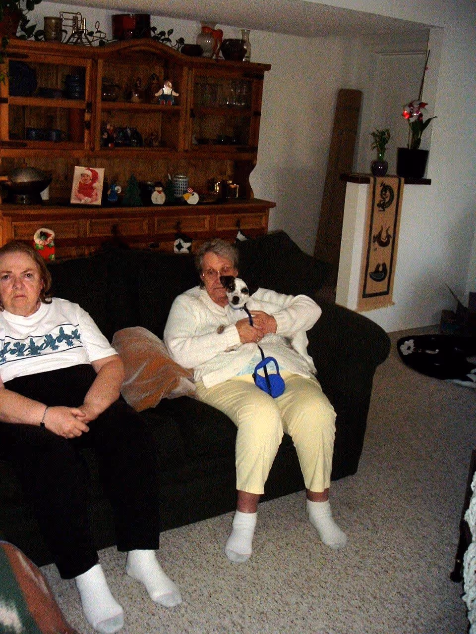 Two elderly women sit on a couch in a living room, one holding a small dog.