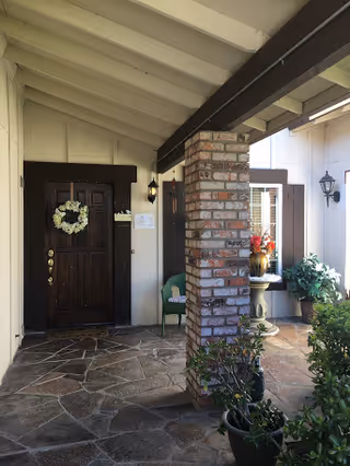 Covered entrance area with a dark wooden door decorated with a white floral wreath. The floor is made of stone tiles, and there is a brick column supporting the roof. A green chair and a small table with a vase of flowers are visible near the window. Potted plants are placed around the entrance.