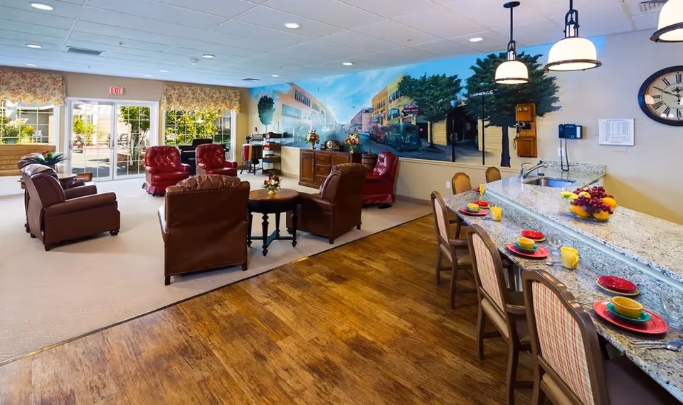 A cozy common area in Canterbury Gardens Memory Care Community featuring a seating area with brown and red leather chairs arranged around a small round table. The room has large windows with floral valances letting in natural light, and a mural on the wall depicting a vintage street scene. Adjacent to the seating area is a granite countertop with chairs set for dining, including colorful plates, bowls, and cups. The floor transitions from wood to carpet, and the room is decorated with flowers and a large wall clock.