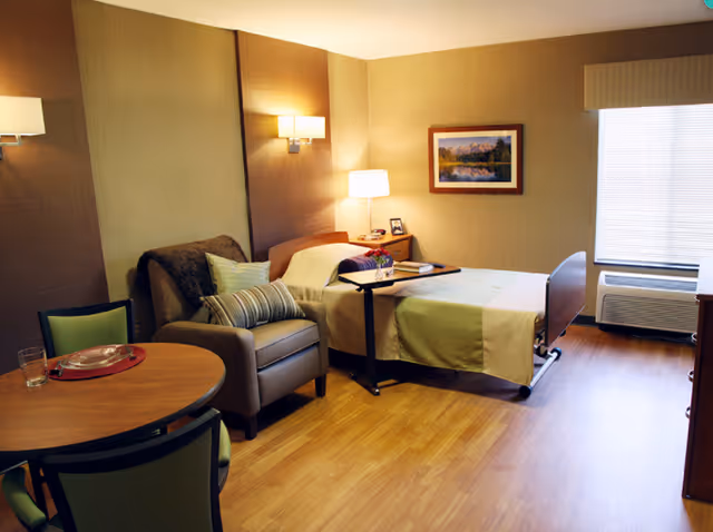A well-lit senior living bedroom with a single bed, a bedside table with a lamp and picture frame, a comfortable armchair with pillows, a round table with chairs, and a large window with blinds. The room has wooden flooring and neutral-colored walls with a framed landscape painting.