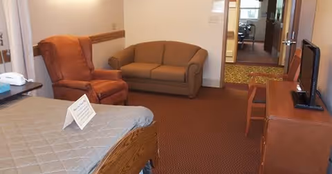 A senior living apartment room with a bed covered in a gray quilt, a brown armchair, a brown loveseat, a wooden dresser with a flat-screen TV on top, and a wooden chair. The room has carpeted flooring and a doorway leading to another area.