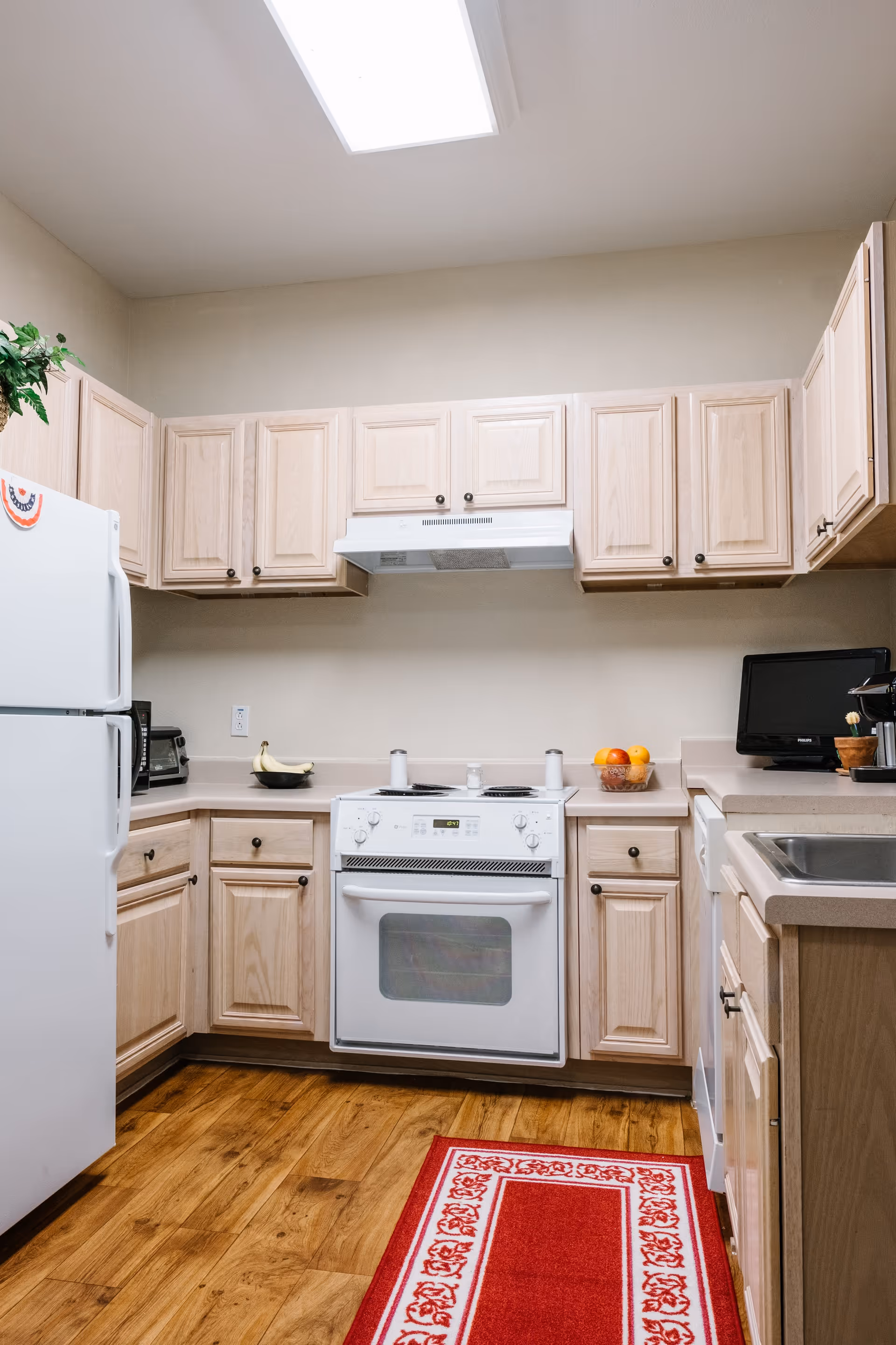 A clean kitchen with light wood cabinets, a white refrigerator, a white oven with stovetop, a microwave, a small television, a coffee maker, a sink, and a red patterned rug on a wooden floor. There is a bowl of bananas and a bowl of oranges on the countertops.