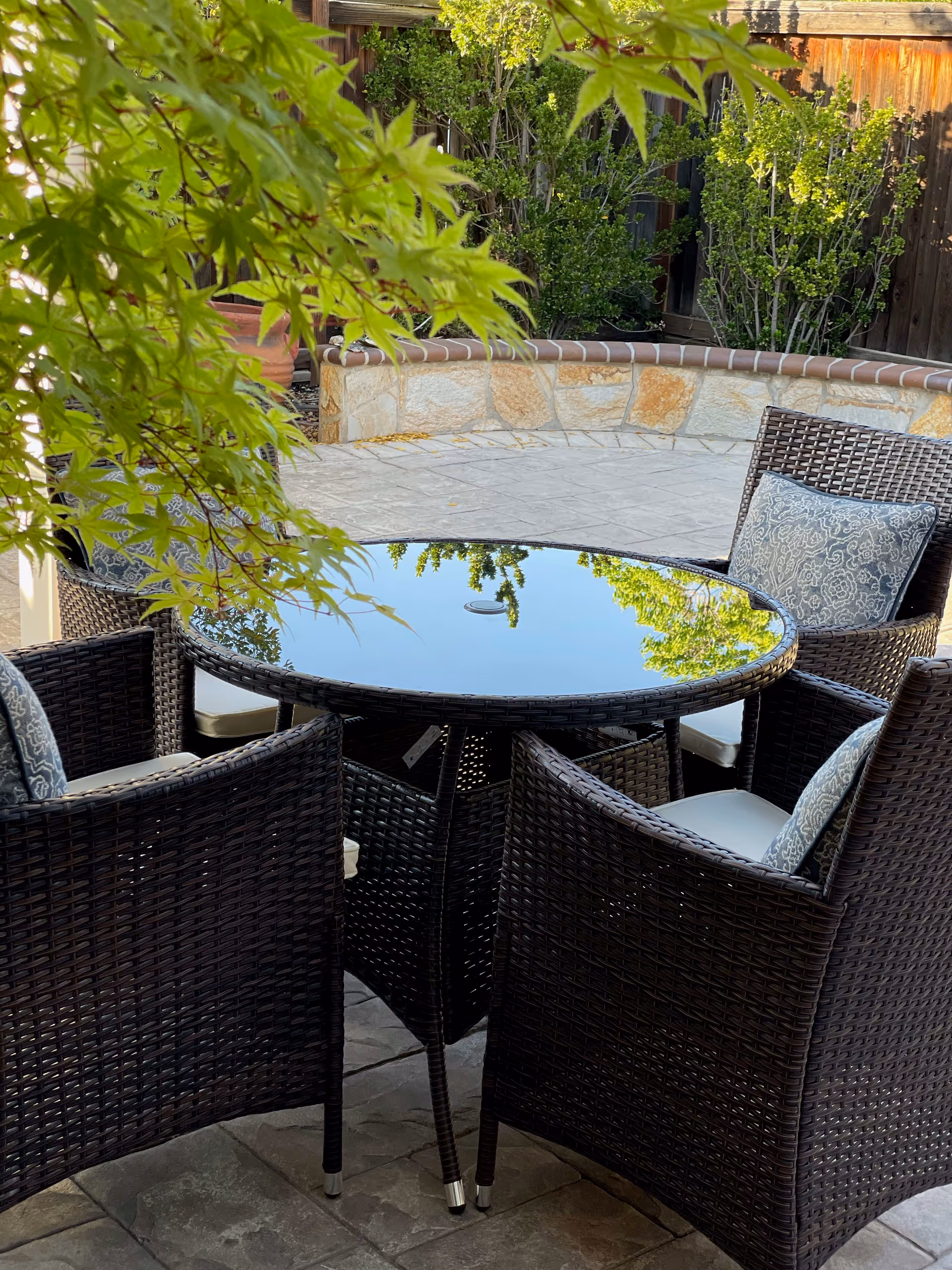 Outdoor patio area with a round glass-top table surrounded by four dark wicker chairs with patterned cushions. Green leafy branches partially frame the scene, and a stone wall with plants is visible in the background.