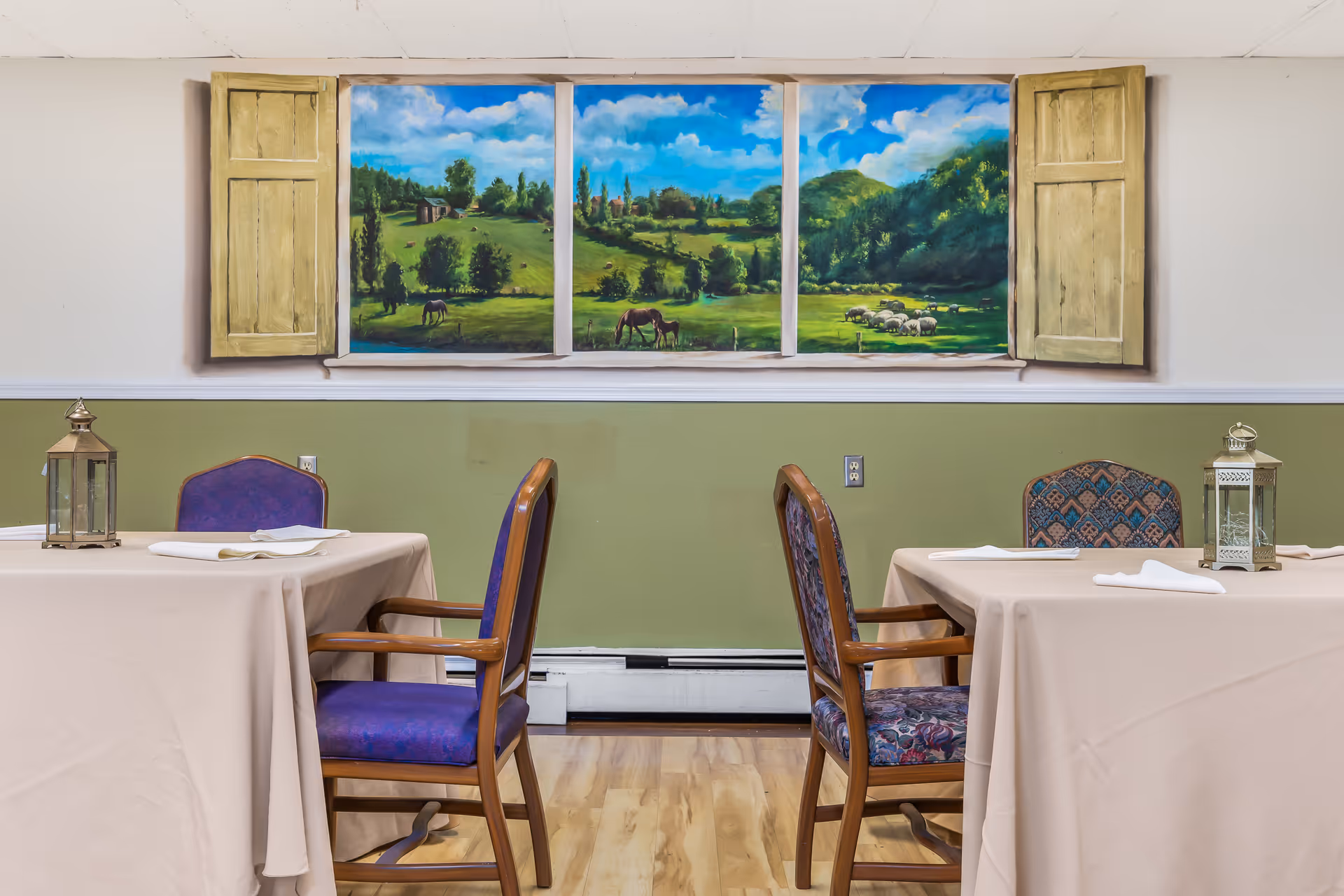 Interior view of a dining room with two tables covered in beige tablecloths, each with two chairs. On each table is a decorative lantern and folded napkins. The wall behind the tables is painted green on the lower half and white on the upper half, featuring a large mural designed to look like a window with open shutters, showing a scenic countryside landscape with horses, sheep, trees, and a blue sky with clouds.