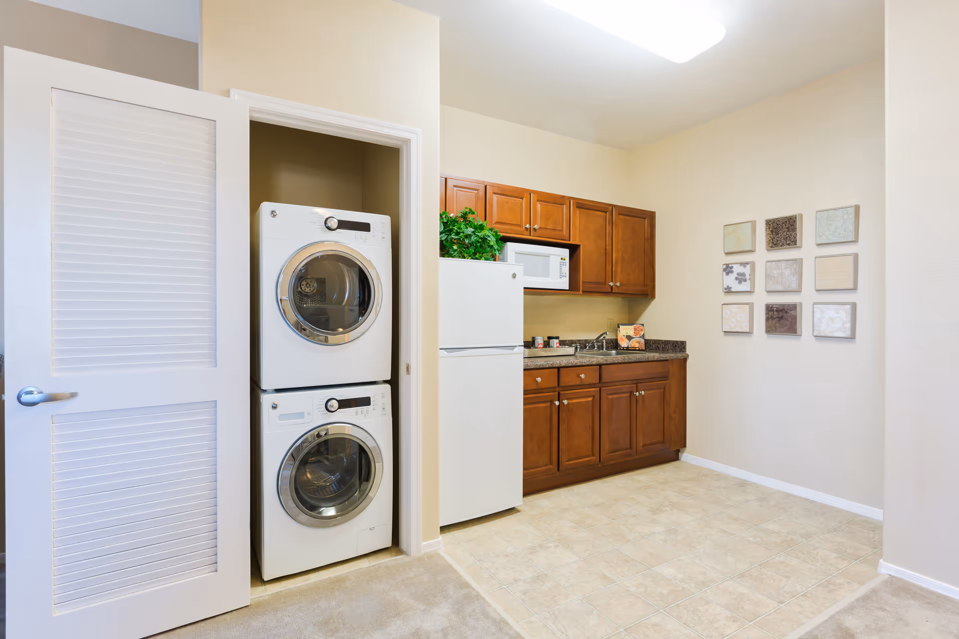 A small kitchen area with wooden cabinets, a white refrigerator, and a microwave. To the left, there is a closet with an open louvered door revealing a stacked washer and dryer. The walls are light-colored, and there are nine decorative square tiles arranged in a grid on the right wall.