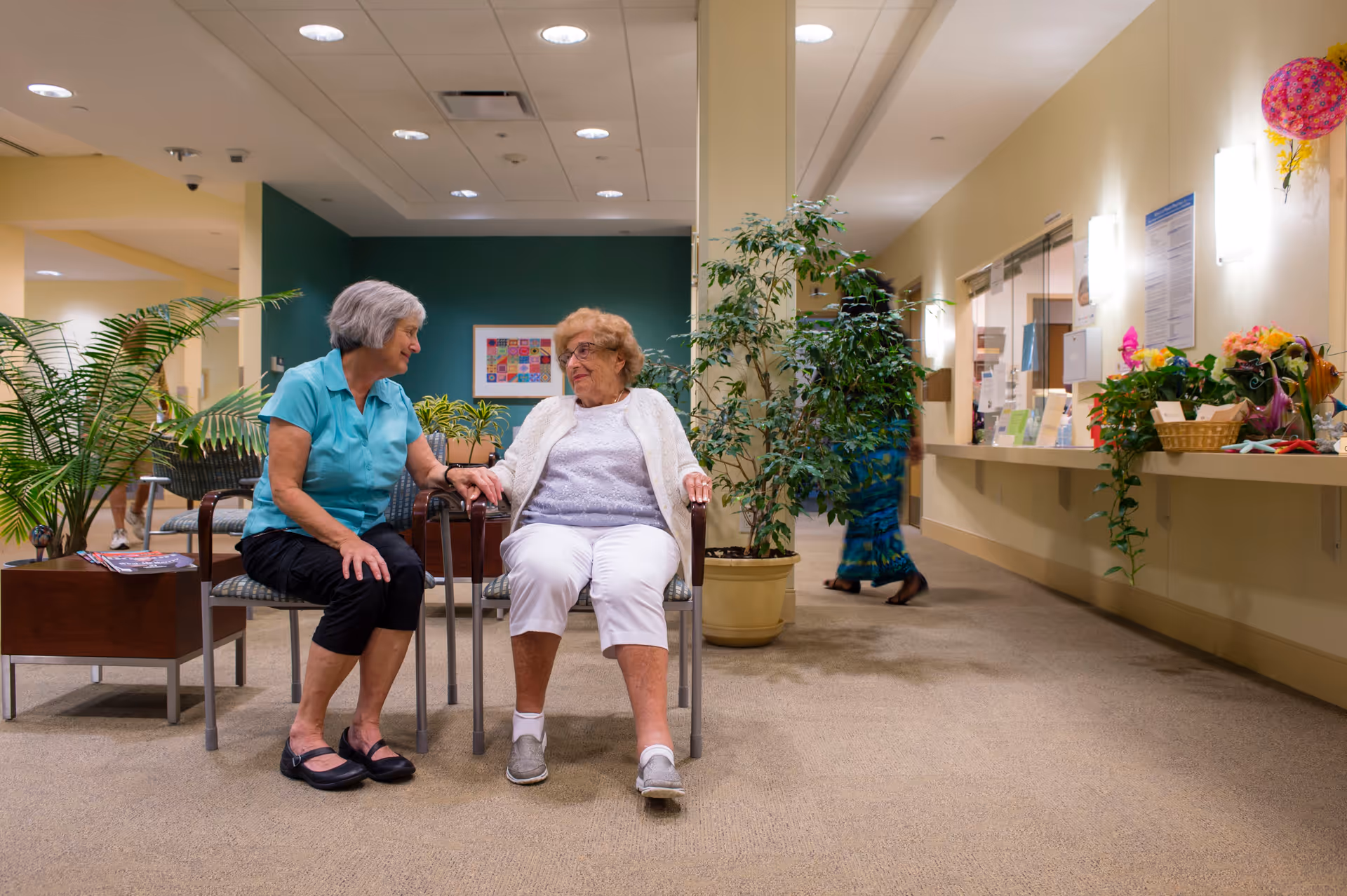 Two elderly women sitting and holding hands in a well-lit indoor common area with plants and a reception desk in the background. One woman is wearing a turquoise shirt and black pants, and the other is wearing a white cardigan and white pants. A person in a blue dress is walking in the background.