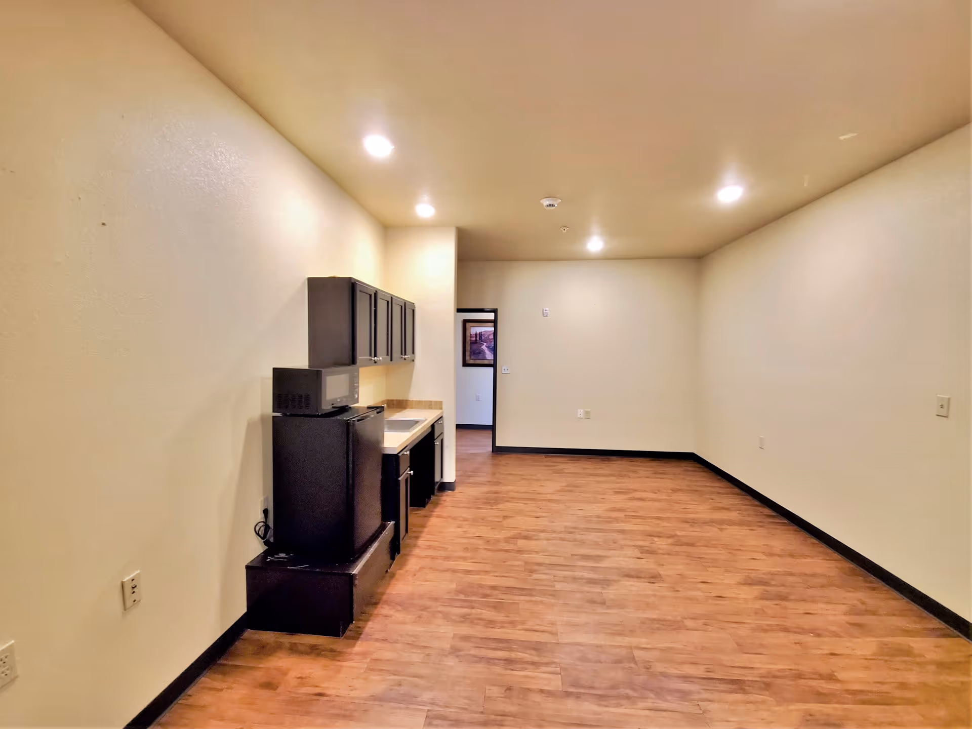 Interior view of a room with wooden flooring and beige walls, featuring a small kitchenette area with dark cabinets, a microwave, and a mini refrigerator. The room is well-lit with recessed ceiling lights and has an open doorway leading to another room.