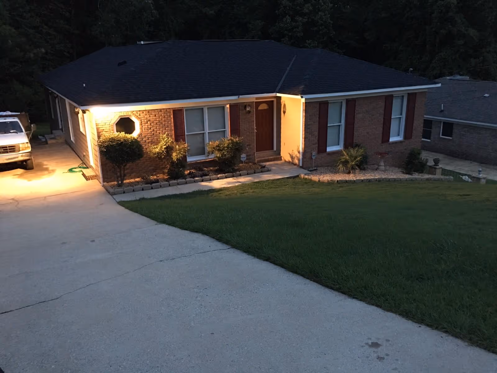 Front exterior of a single-story brick house with a lit entry, driveway, and grassy front yard at dusk.