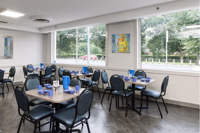 A bright dining room with several tables and chairs arranged neatly. Each table is set with blue cups, bowls, and pitchers. Large windows allow natural light to fill the room and provide a view of green trees outside. The walls are decorated with colorful abstract paintings.
