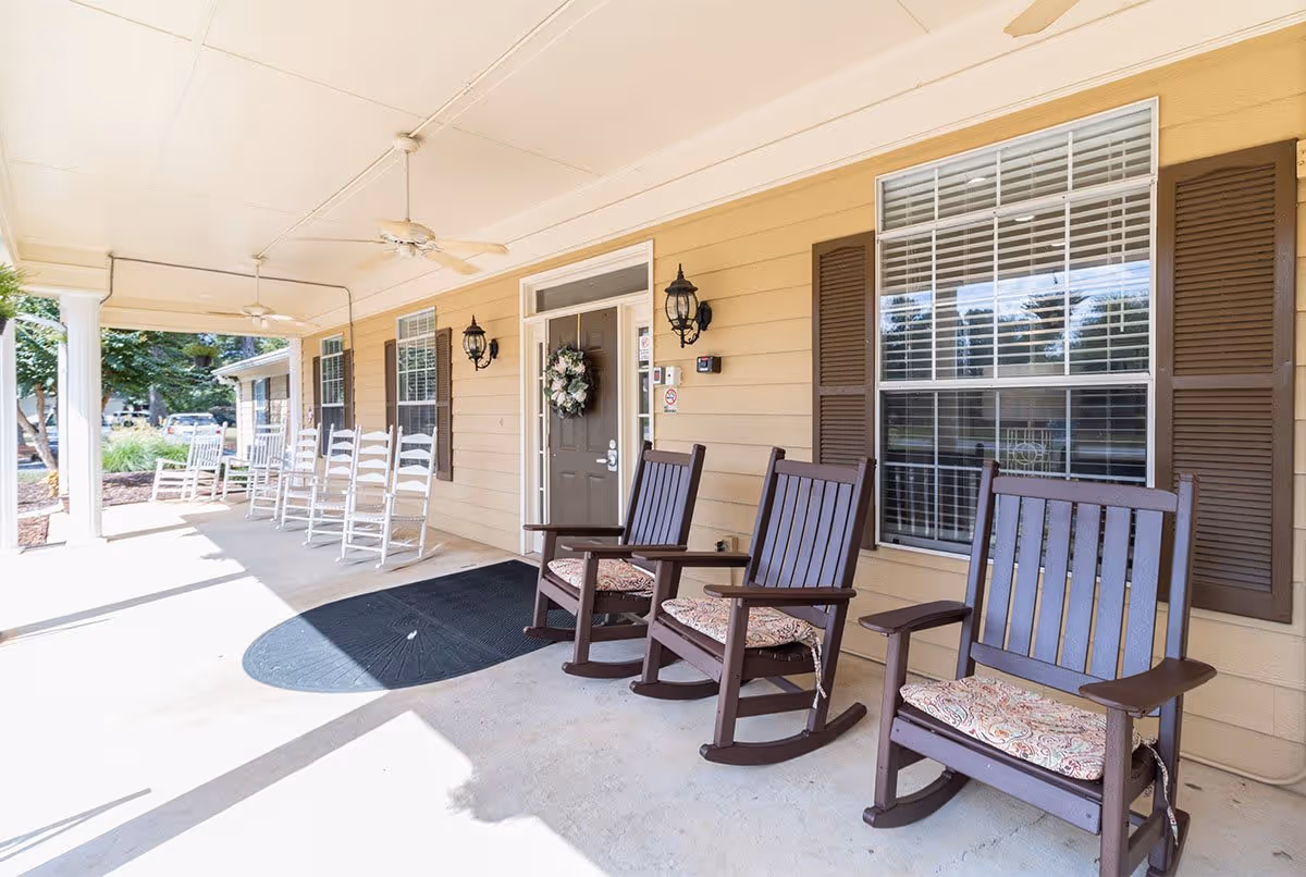A covered porch area at Tipton Place with several rocking chairs, including three dark wooden rocking chairs with patterned cushions and multiple white rocking chairs lined up along the wall. The porch has ceiling fans and is adjacent to a beige building with brown shutters and windows.