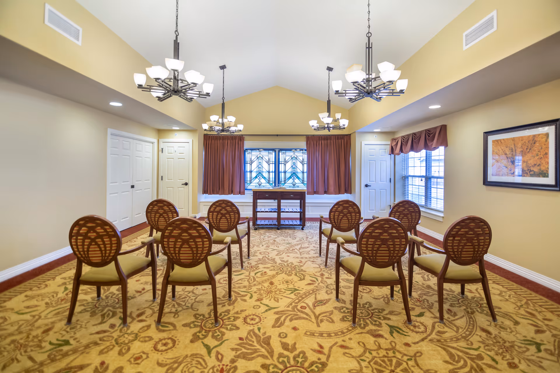 A small meeting or activity room with eight wooden chairs arranged in two rows facing a window with stained glass and brown curtains. The room has beige walls, a patterned carpet, three chandeliers hanging from a vaulted ceiling, and a framed picture on the right wall.
