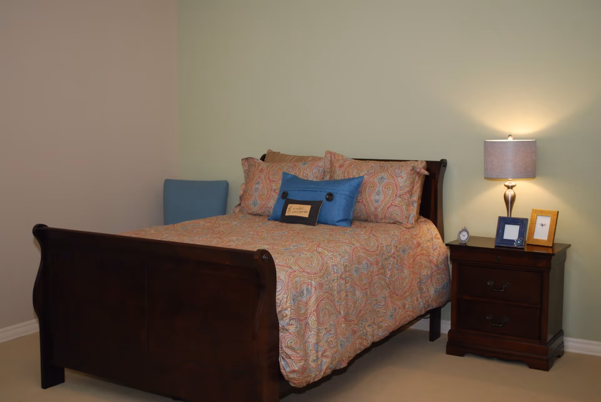 A bedroom with a wooden bed frame and a paisley-patterned bedspread in shades of orange and pink. The bed has multiple pillows, including a blue decorative pillow. Next to the bed is a wooden nightstand with two drawers, a table lamp with a beige shade, a small clock, and two picture frames. The walls are painted in light colors, one beige and one light green.