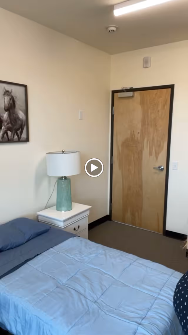 A simple bedroom with a bed covered in a light blue comforter and dark blue pillow. Next to the bed is a white nightstand with a greenish-blue lamp on it. A wooden door with a silver handle is visible in the background, along with a framed picture of horses on the wall.