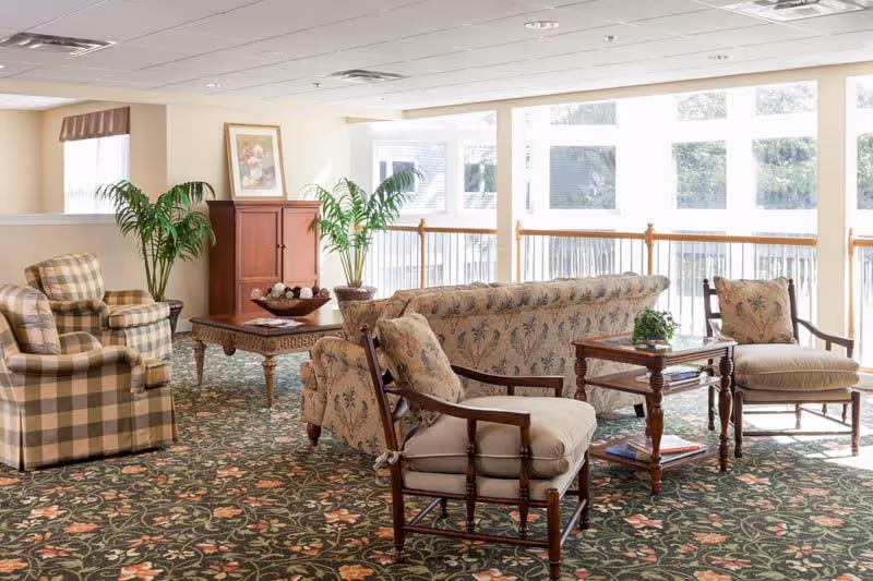A bright and spacious living room area with large windows allowing natural light to fill the space. The room features a floral patterned carpet, a floral upholstered sofa, two cushioned armchairs with wooden frames, a plaid armchair, a wooden coffee table with decorative items, a side table with a small plant, and two potted plants near a wooden cabinet with a framed picture above it.