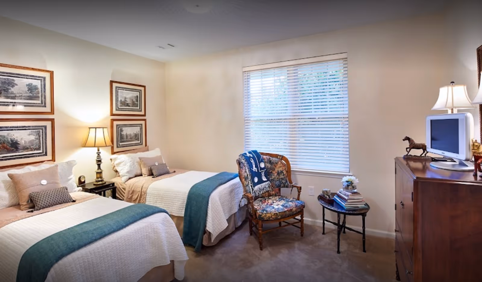 A bedroom with two twin beds, each made with white quilts and teal throws. Between the beds is a small nightstand with a lamp. On the wall above the beds are four framed pictures. A floral upholstered chair with a blue blanket draped over it sits near a window with white blinds. Next to the chair is a small table stacked with books and a small flower arrangement. A wooden dresser with a small TV and decorative horse figurines is on the right side of the room.