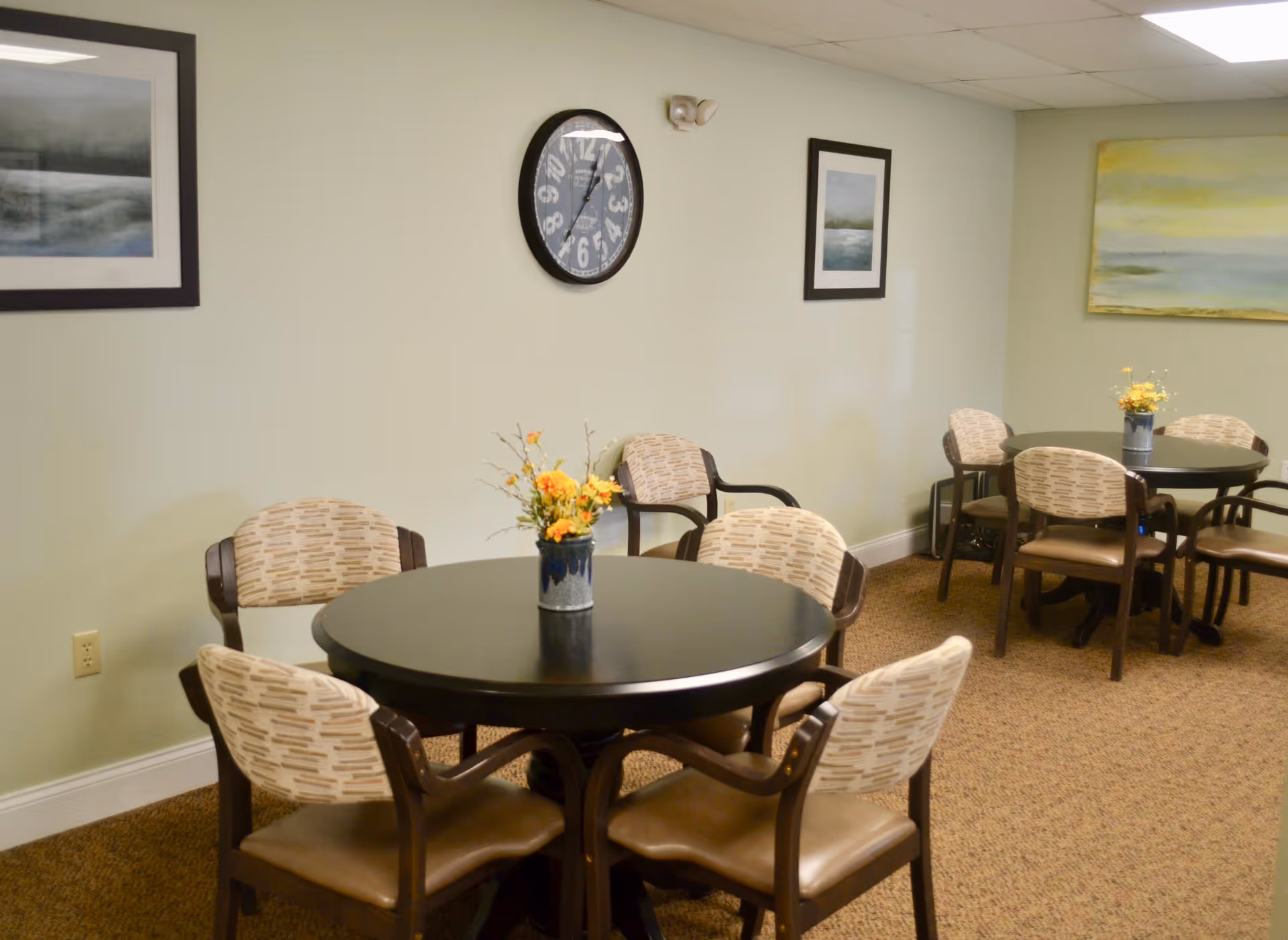 A senior living facility common dining area with two round black tables, each surrounded by four cushioned chairs with patterned backs and brown seats. Each table has a small vase with yellow and orange flowers. The walls are light green with framed landscape paintings and a large round clock showing 12:05. The floor is carpeted in a brown pattern.