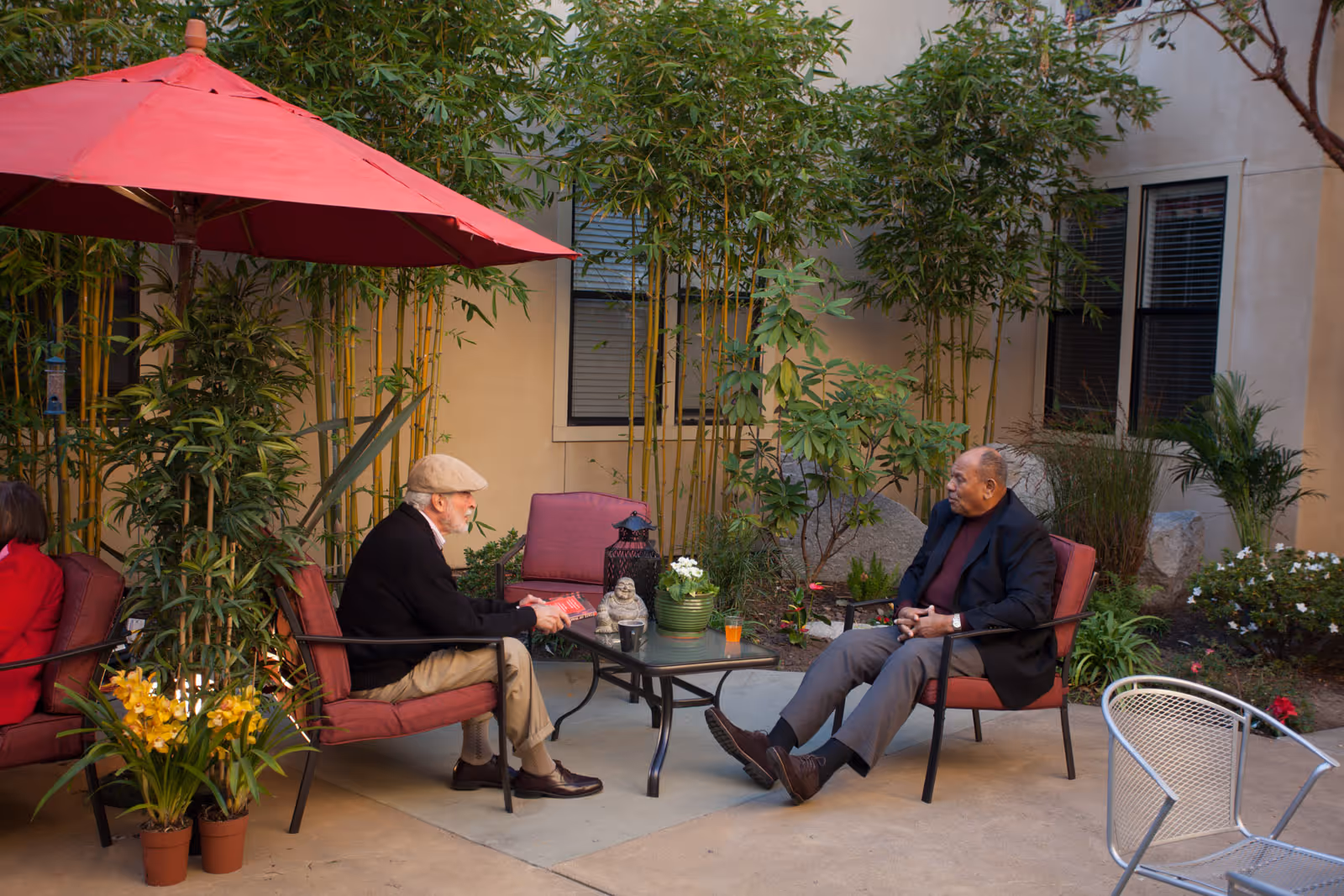 Two elderly men sit and talk in a landscaped courtyard patio with a red umbrella, potted plants, and patio chairs.