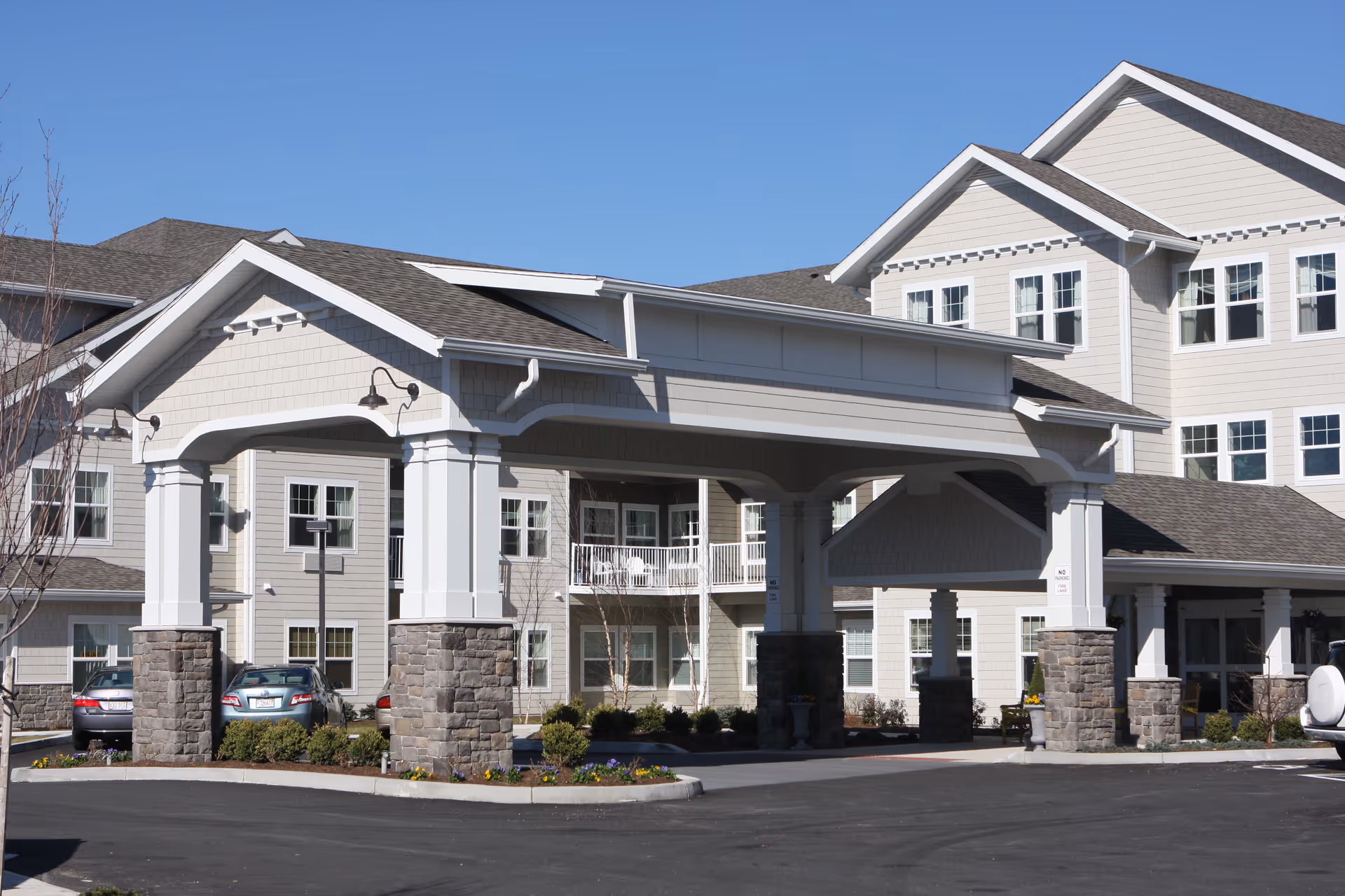 Exterior view of a multi-story senior living facility building with beige siding and white trim. The entrance features a large covered porte-cochère supported by stone and white columns. Several windows are visible, along with a few parked cars and some landscaping with small bushes and flowers. The sky is clear and blue.