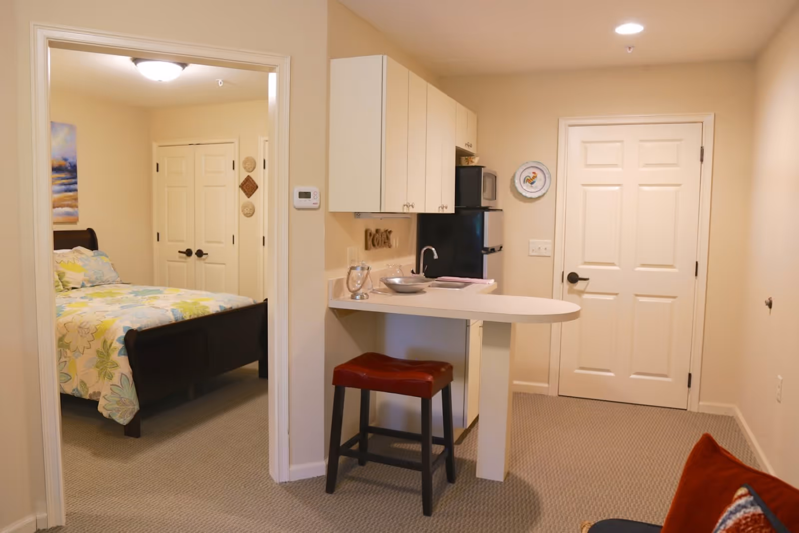 Interior view of a senior living facility room showing a small kitchenette with white cabinets, a mini fridge, microwave, and a counter with a red cushioned stool. To the left, there is an open doorway leading to a bedroom with a bed covered in floral bedding. The walls are painted beige and the floor is carpeted.