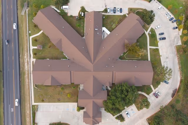 Aerial view of a large building with a unique star-shaped roof design, surrounded by parking lots, trees, and adjacent to a road with vehicles.