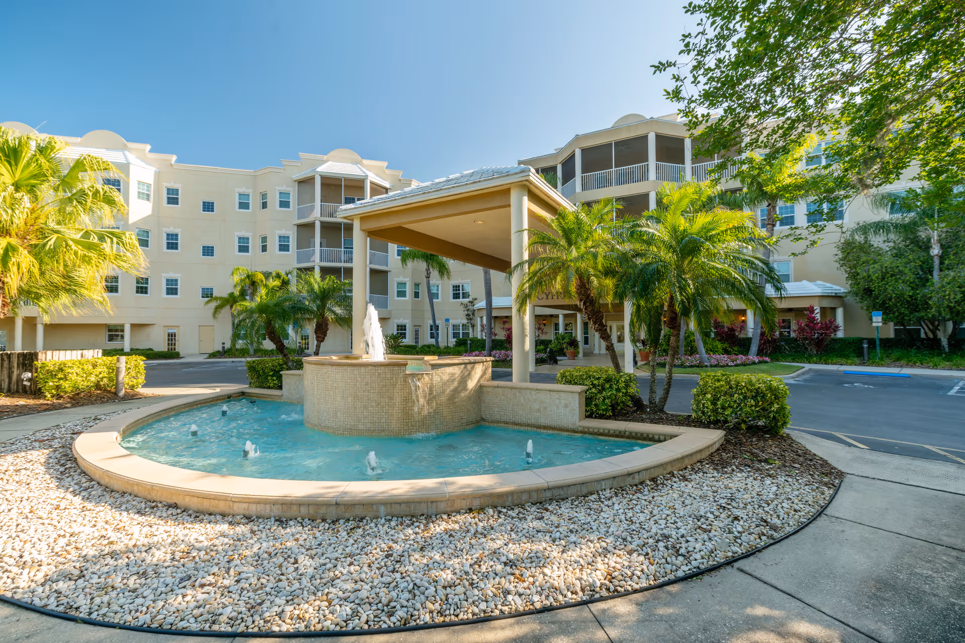 Front entrance of a multi-story beige building with a covered porte-cochere, a circular fountain, palm trees, and landscaped grounds.