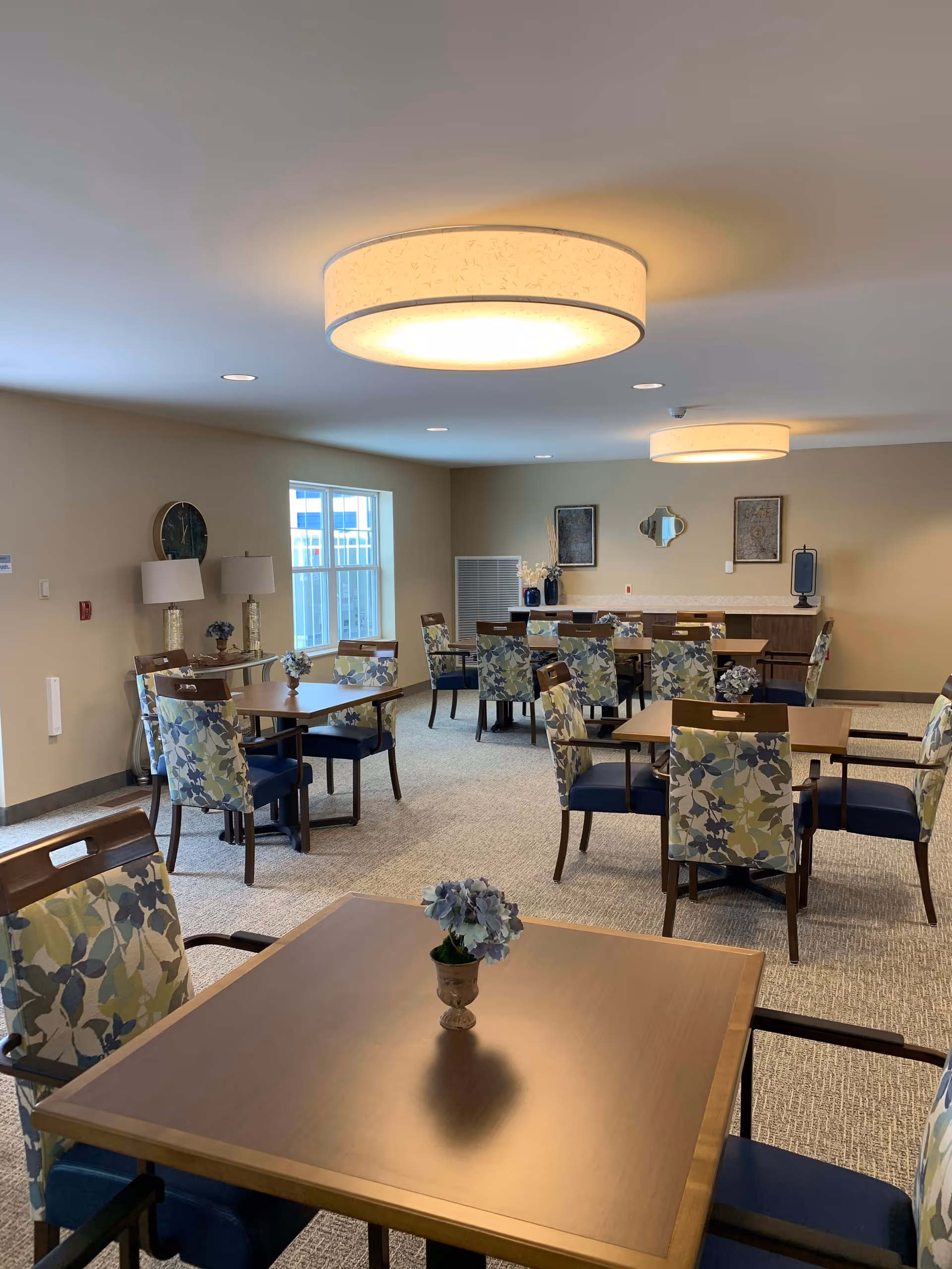 A well-lit dining room with several wooden tables and chairs featuring floral-patterned upholstery. Each table has a small vase with flowers. The room has beige walls, carpeted floor, two large circular ceiling lights, and windows letting in natural light. Decorative items and framed artwork are on the far wall.