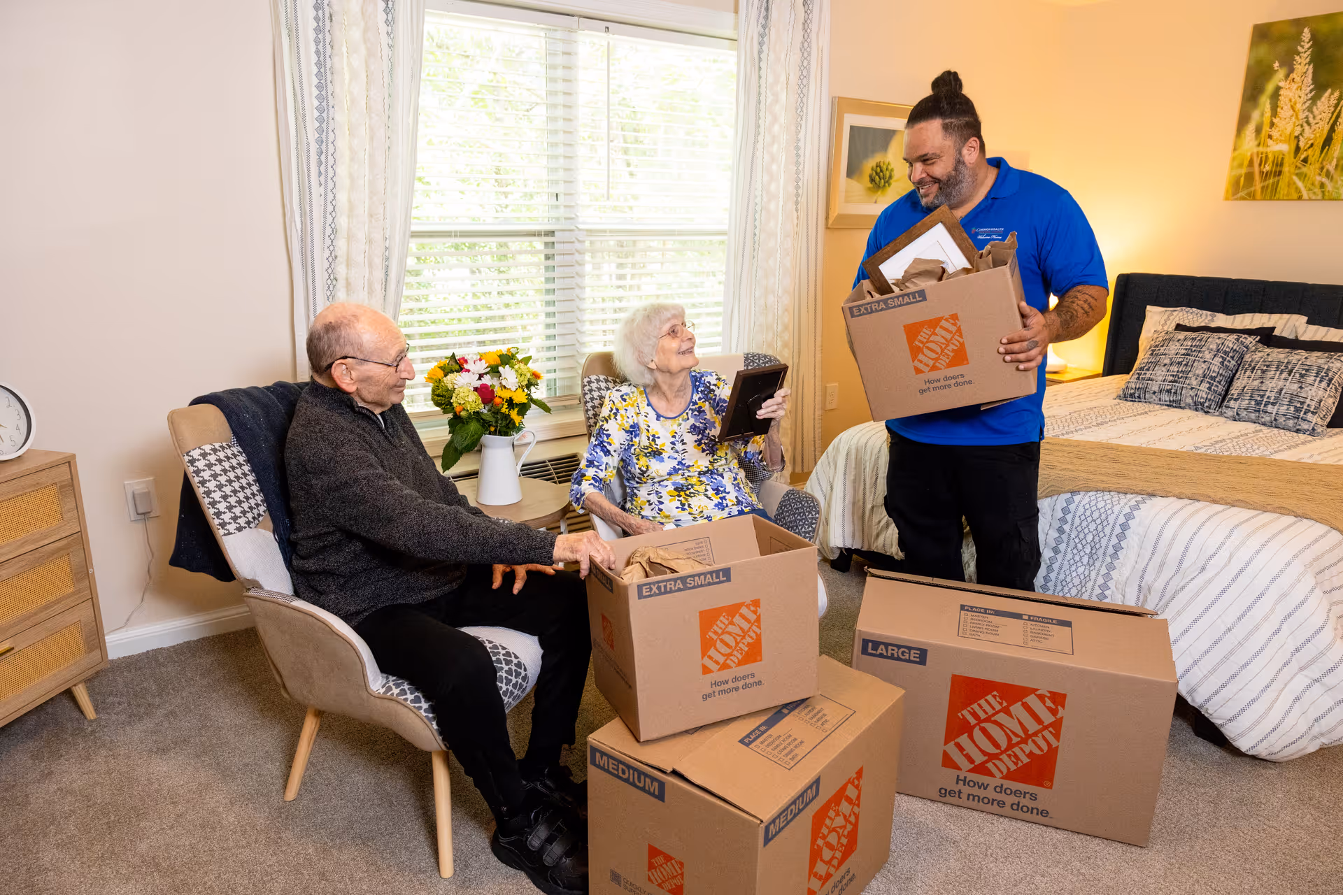 An elderly man and woman sitting in a cozy bedroom with a younger man standing nearby holding a moving box. The elderly woman is holding a framed photo and smiling at the younger man. Several moving boxes labeled 'The Home Depot' are stacked in the room. The bedroom has a bed with patterned bedding, a nightstand with a lamp, and a window with curtains letting in natural light.