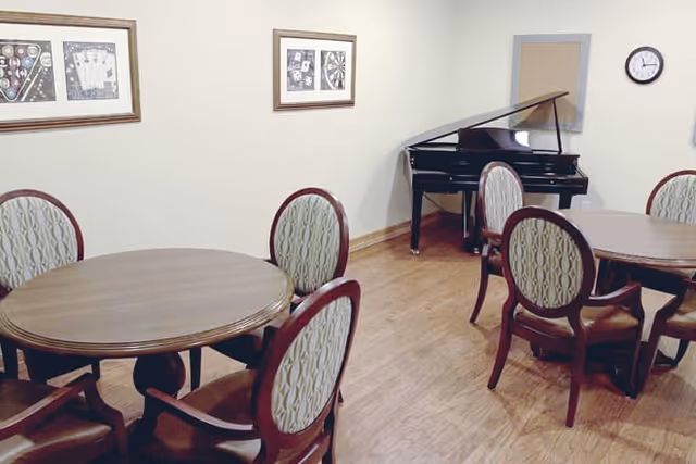 A room with round wooden tables and upholstered chairs arranged around them. A black grand piano is positioned against the far wall. The walls are decorated with framed artwork and a clock is mounted on the wall.