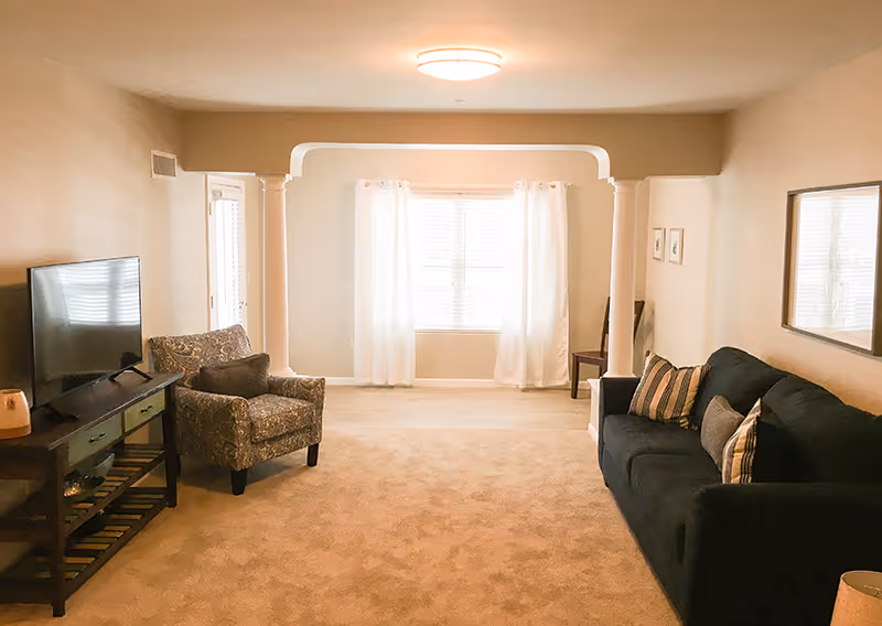 A bright living room with beige walls and carpet. The room features a dark blue sofa with striped and solid pillows on the right, a patterned armchair on the left, and a TV on a wooden stand. There are two white columns framing a window with white curtains at the far end of the room, and a ceiling light fixture above.