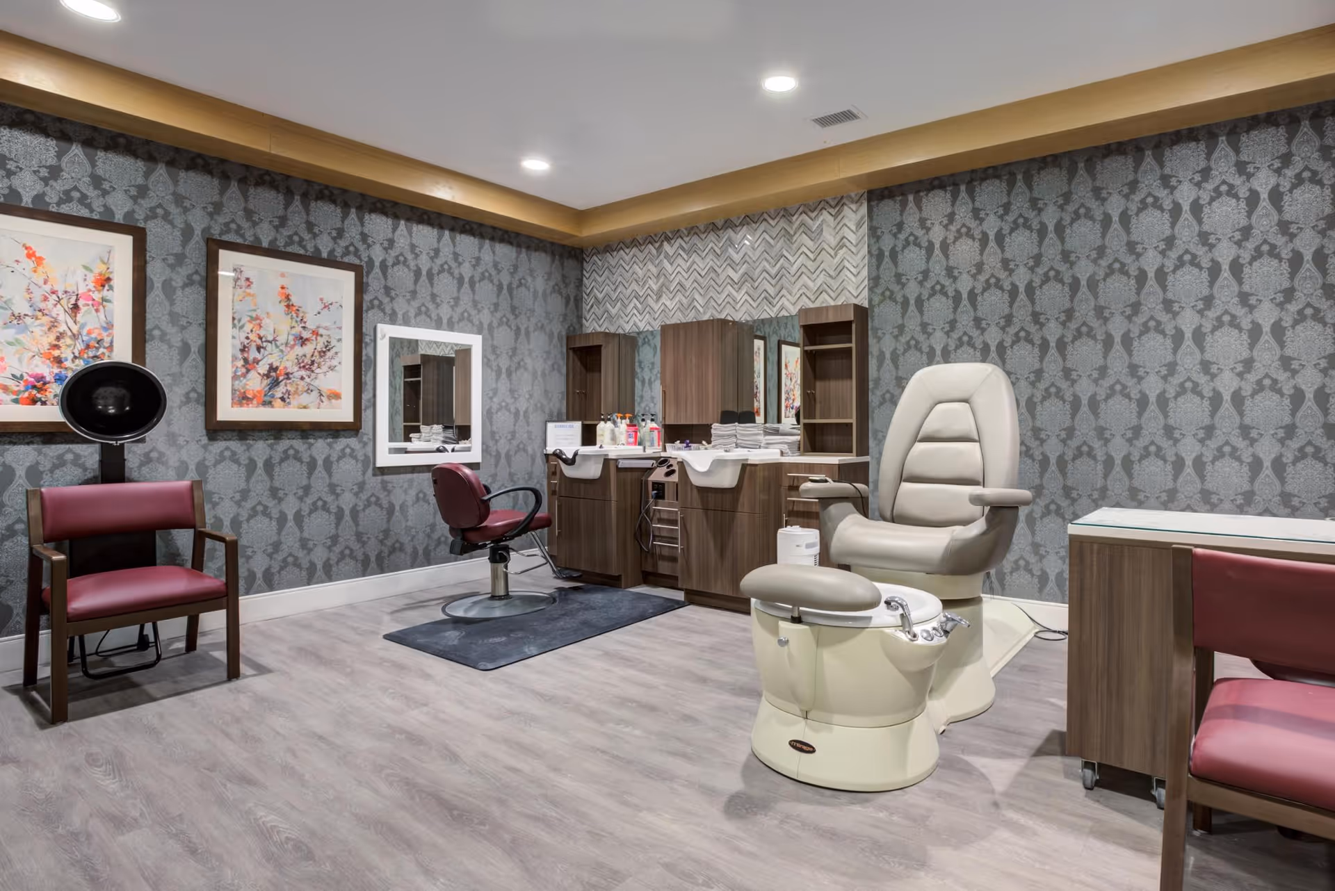 Interior of a salon room with a pedicure chair, a hair washing station with a salon chair, two framed floral artworks on the wall, a hair dryer chair, and patterned wallpaper.