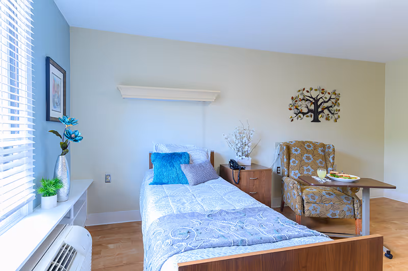 A bright and cozy senior living bedroom with a single bed made up with white and blue bedding and pillows. Next to the bed is a wooden nightstand with a telephone and a decorative plant. A floral patterned armchair sits beside a small table with a meal and a glass of juice. The room has light-colored walls, a window with blinds, and decorative wall art above the chair.