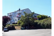 Exterior view of a multi-story residential building with balconies, surrounded by trees and bushes, with a parking area in the foreground containing a few cars and a dumpster.