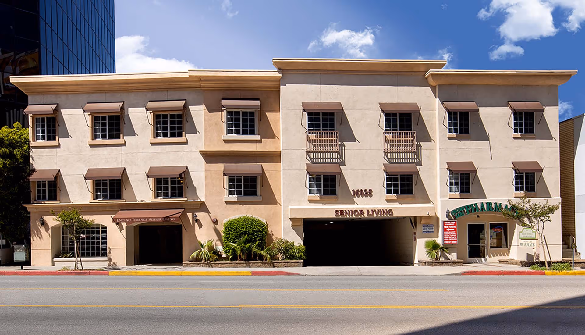 Exterior front view of Encino Terrace Senior Living building with beige walls, multiple windows with brown awnings, a driveway entrance, and a pharmacy on the right side under a green sign.