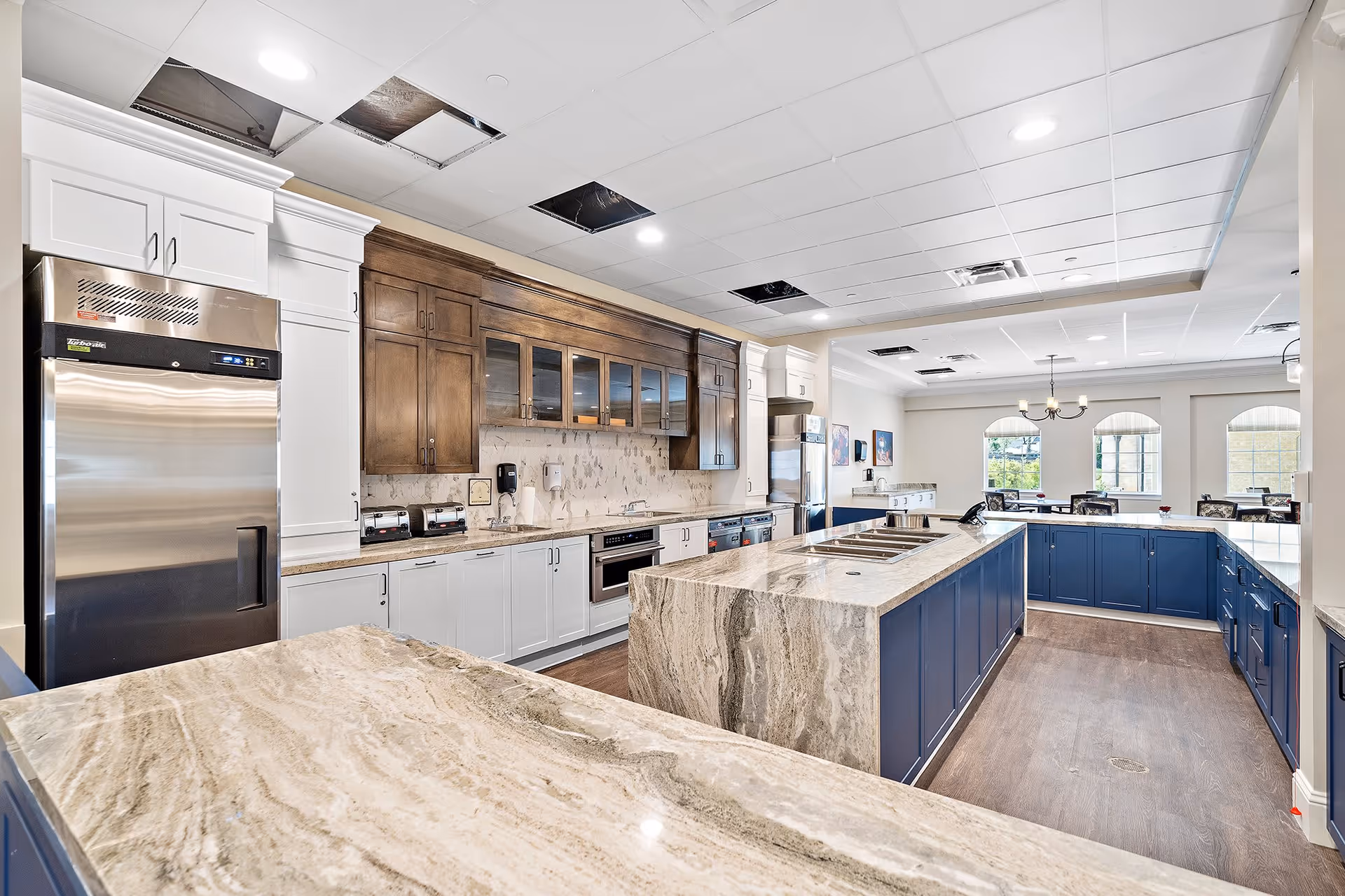 A spacious, modern kitchen with marble countertops and a large island in the center. The kitchen features white and dark wood cabinetry, stainless steel appliances including a refrigerator and oven, and a dining area with tables and chairs visible in the background. The ceiling has recessed lighting and some open panels.