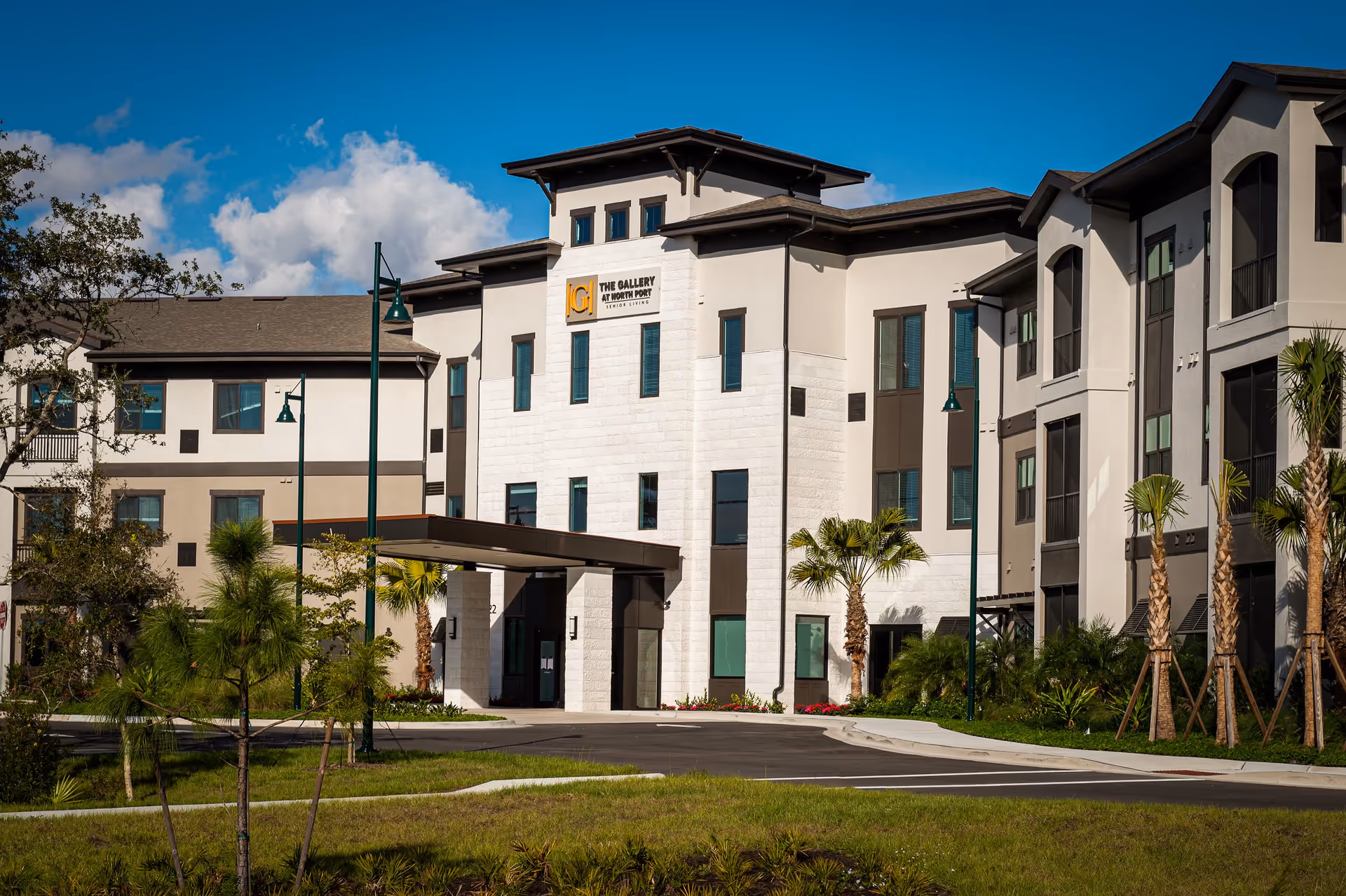 Exterior view of The Gallery at North Port senior living facility building with a driveway, palm trees, and green landscaping under a blue sky with some clouds.