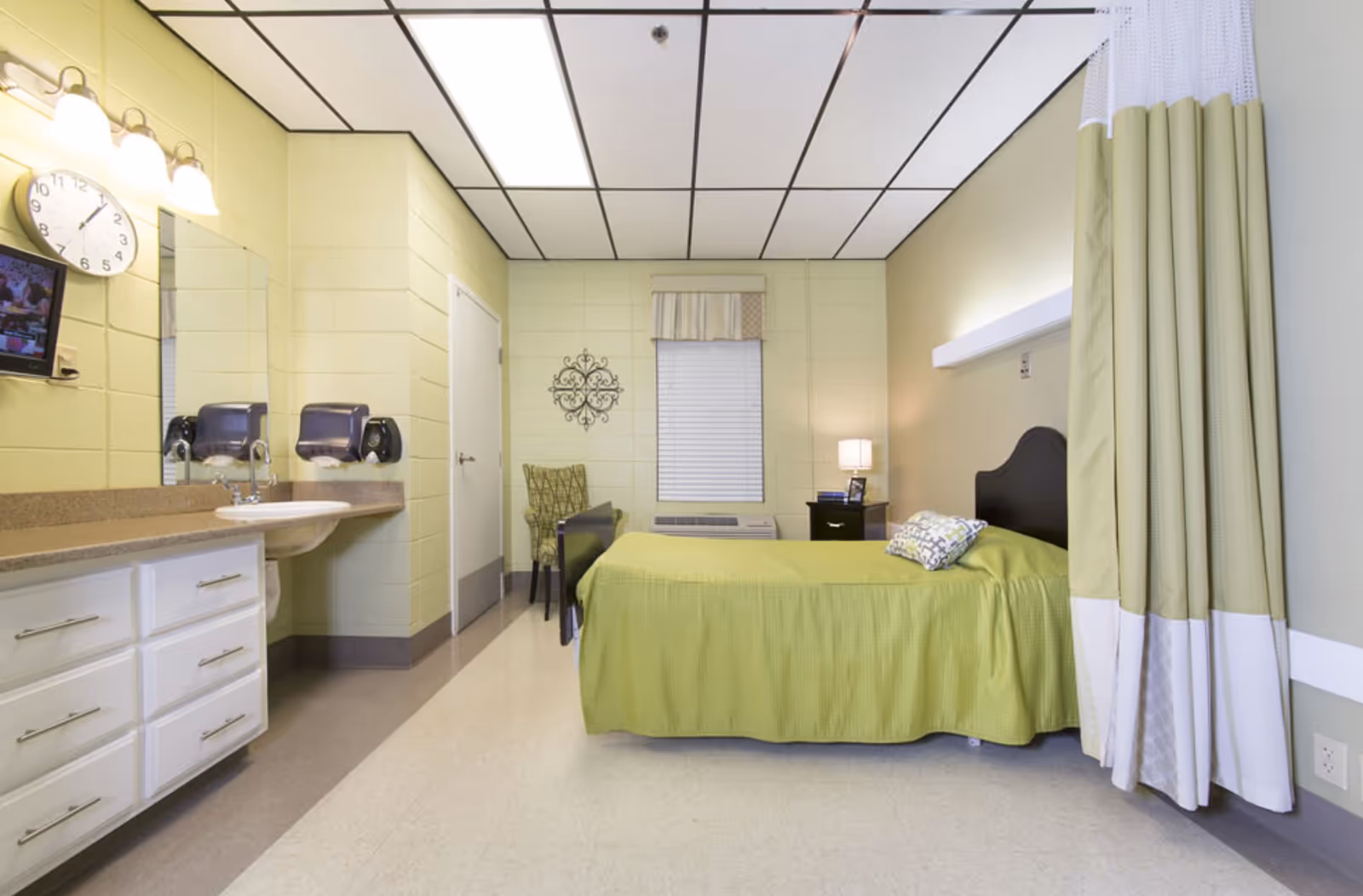 A simple, clean bedroom in a senior living facility with a single bed covered in a green bedspread and a patterned pillow. The room has a window with blinds and a valance, a nightstand with a lamp and picture frame, a green patterned chair, and a wall-mounted sink with a mirror and soap dispensers. There is a clock on the wall and a privacy curtain partially drawn on the right side.
