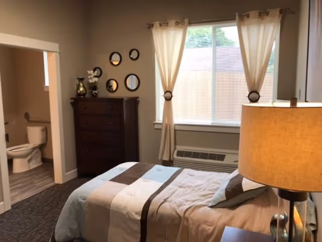 A bedroom with a bed covered in a beige, brown, and blue striped comforter. There is a wooden dresser with decorative items and round mirrors on the wall above it. A window with beige curtains lets in natural light. To the left, an open door reveals a bathroom with a toilet and a window.