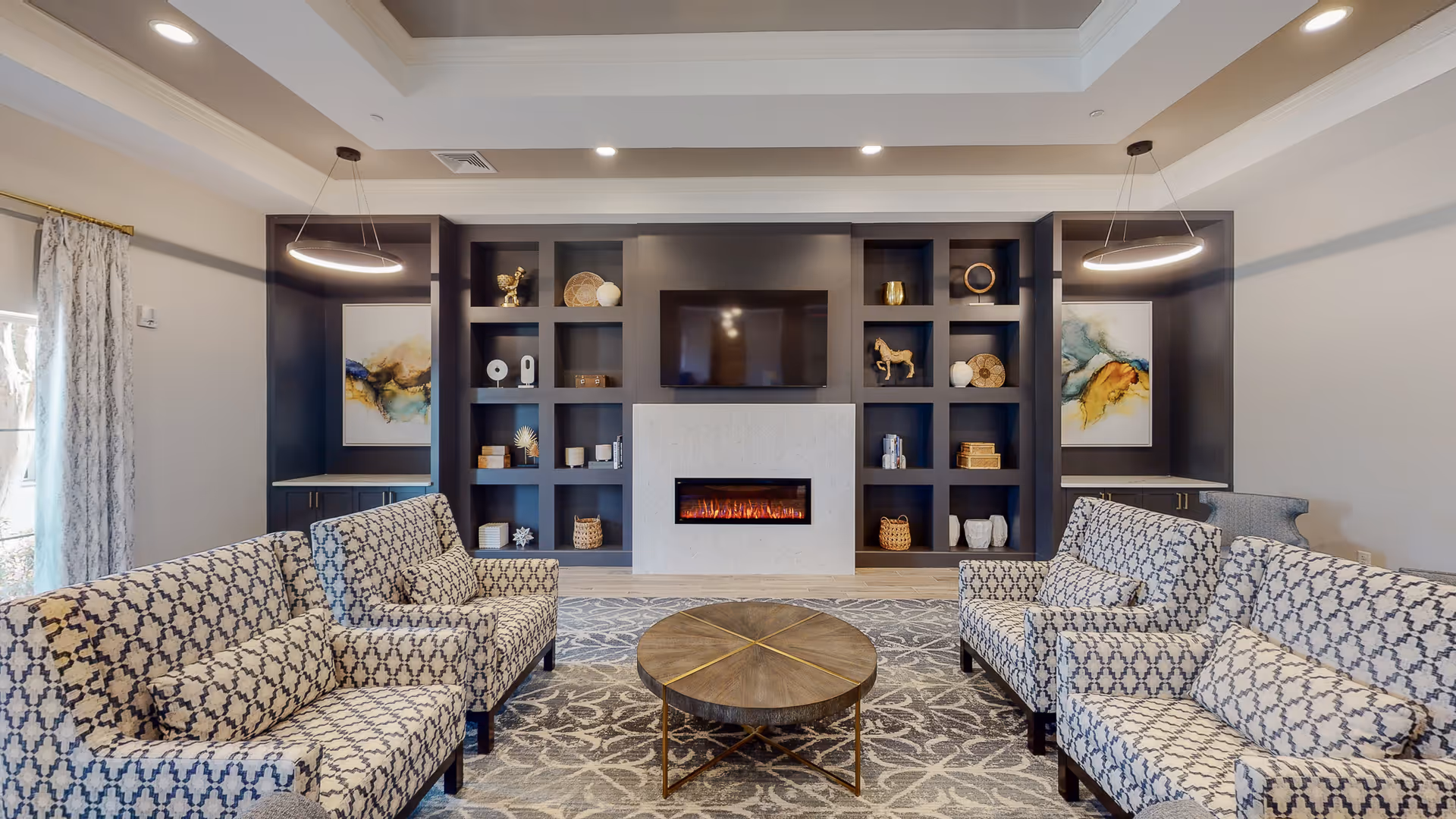 Communal lounge with patterned sofas around a round coffee table facing a wall-mounted TV and electric fireplace with built-in shelving.