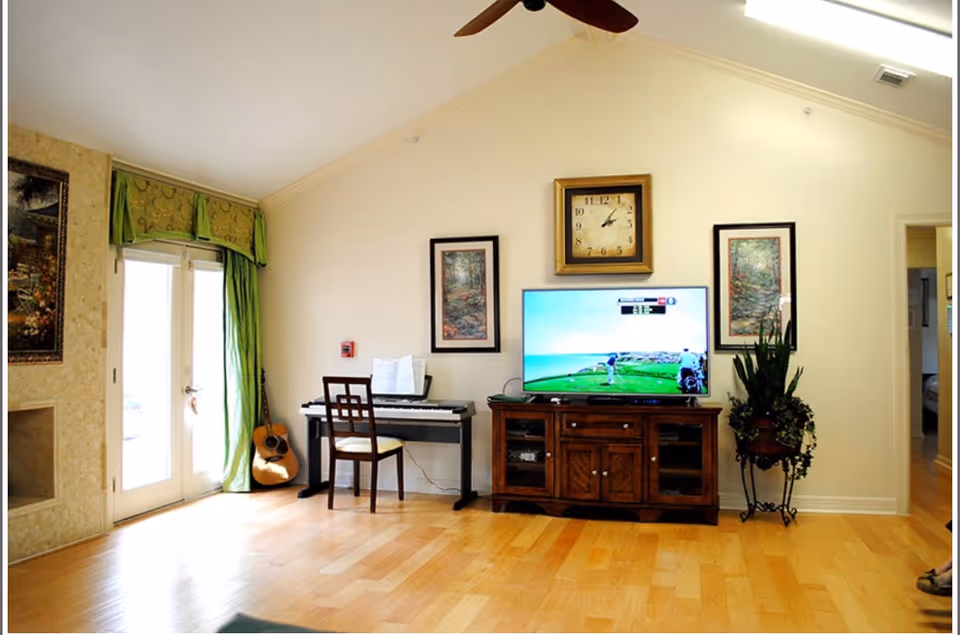 A bright living room with light wooden flooring, a digital piano with a chair, a guitar leaning against the wall, a wooden TV stand with a flat-screen TV displaying a golf game, two framed pictures on the wall, a large clock above the TV, a potted plant on a stand, and French doors with green curtains letting in natural light.