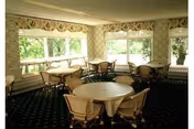 A dining room with round tables covered with white tablecloths and surrounded by chairs. Large windows with floral valances allow natural light to fill the room, and the carpet has a dark pattern.