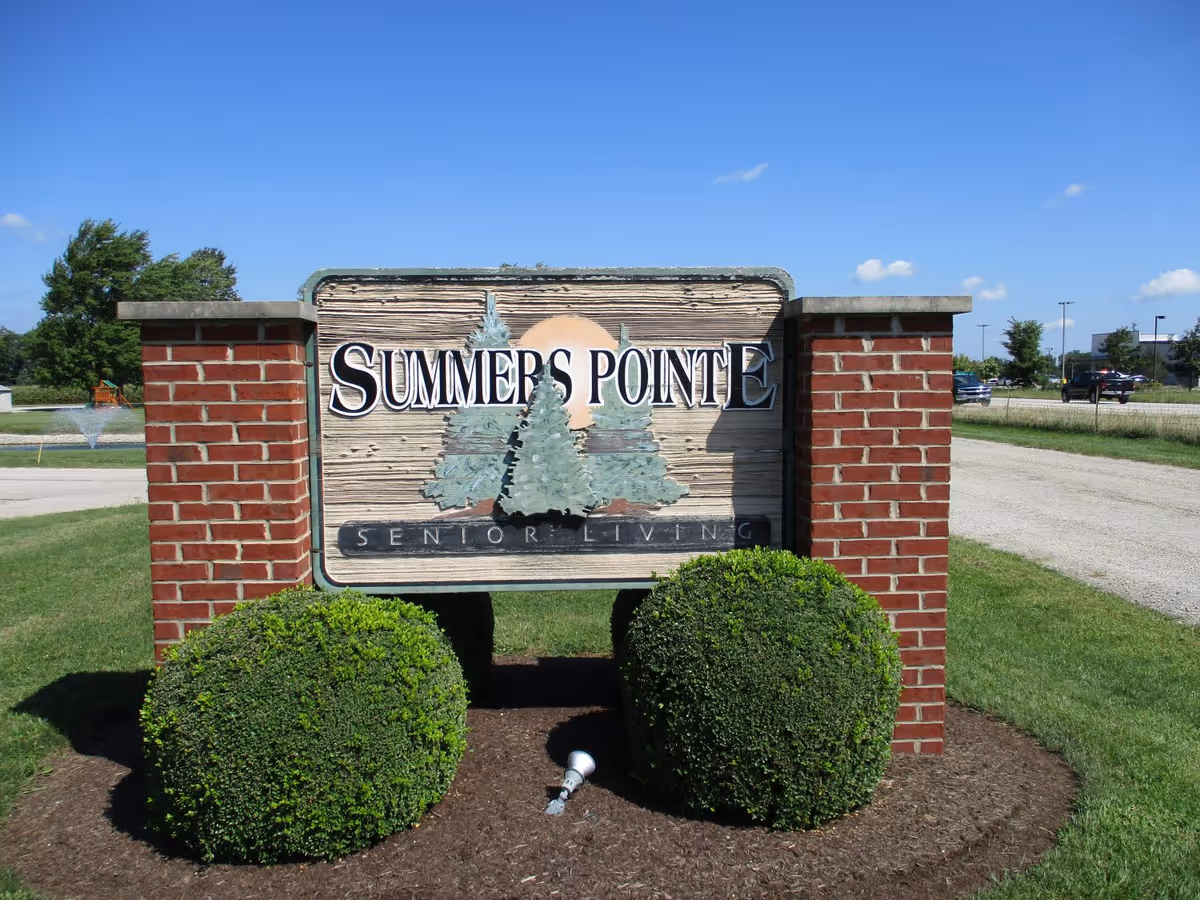 Outdoor sign for Summers Pointe Senior Living mounted on a wooden board between two brick pillars with two neatly trimmed bushes in front, set against a clear blue sky and grassy area.