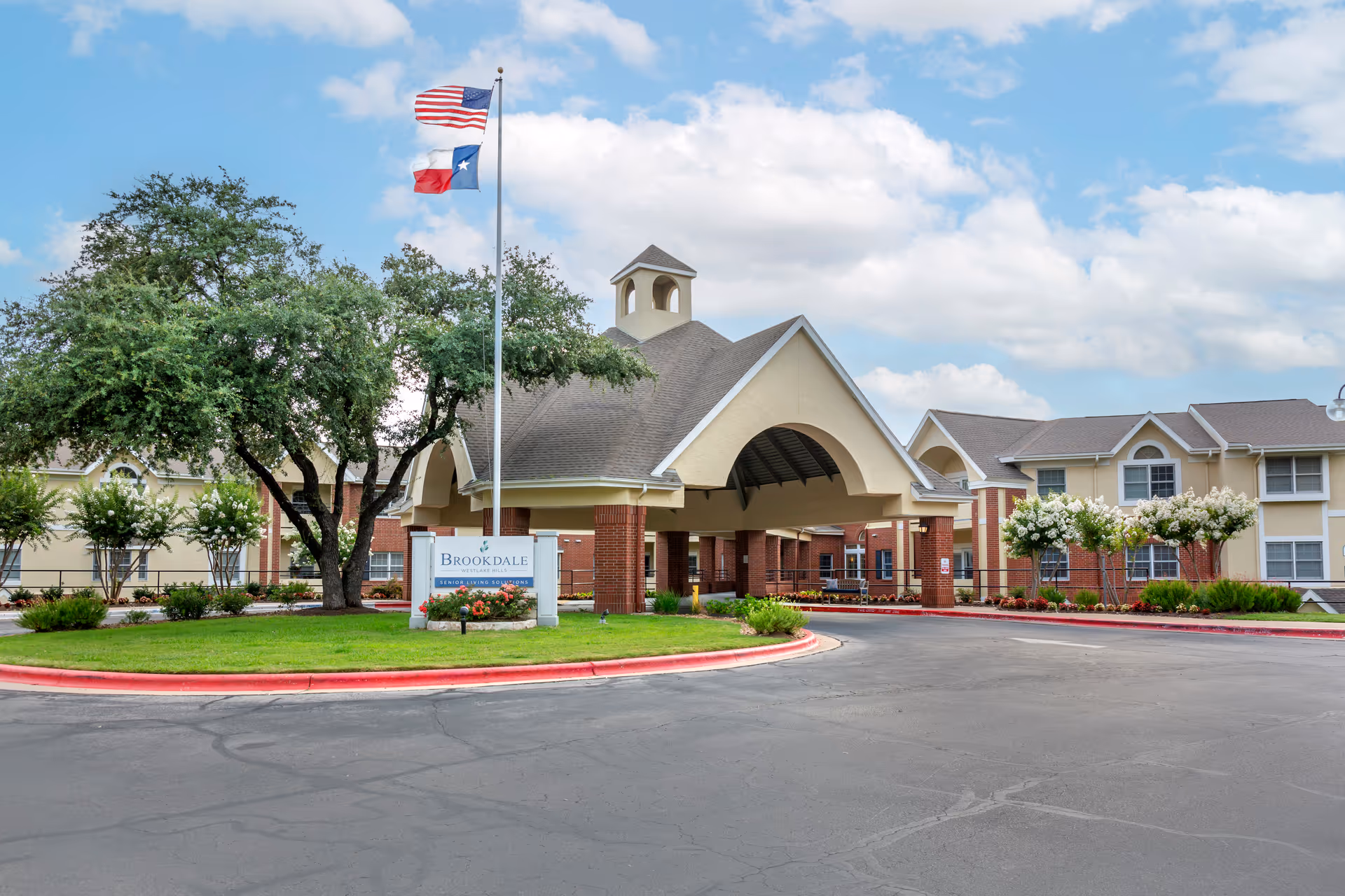 Front entrance and porte-cochère of Brookdale Westlake Hills senior living building with American and Texas flags and surrounding landscaping.