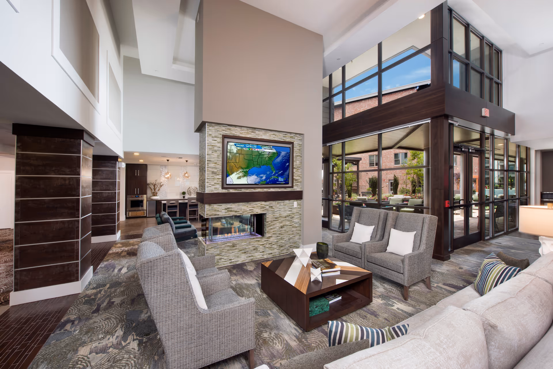 A modern living room area in a senior living facility featuring a stone fireplace with a mounted flat-screen TV above it, several gray upholstered armchairs and a sofa with striped pillows, a wooden coffee table with decorative items, large windows providing natural light, and a view of an outdoor patio area.