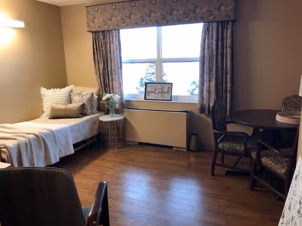 A furnished senior living bedroom with a single bed by a window, a side table with flowers, and a small dining table with chairs.