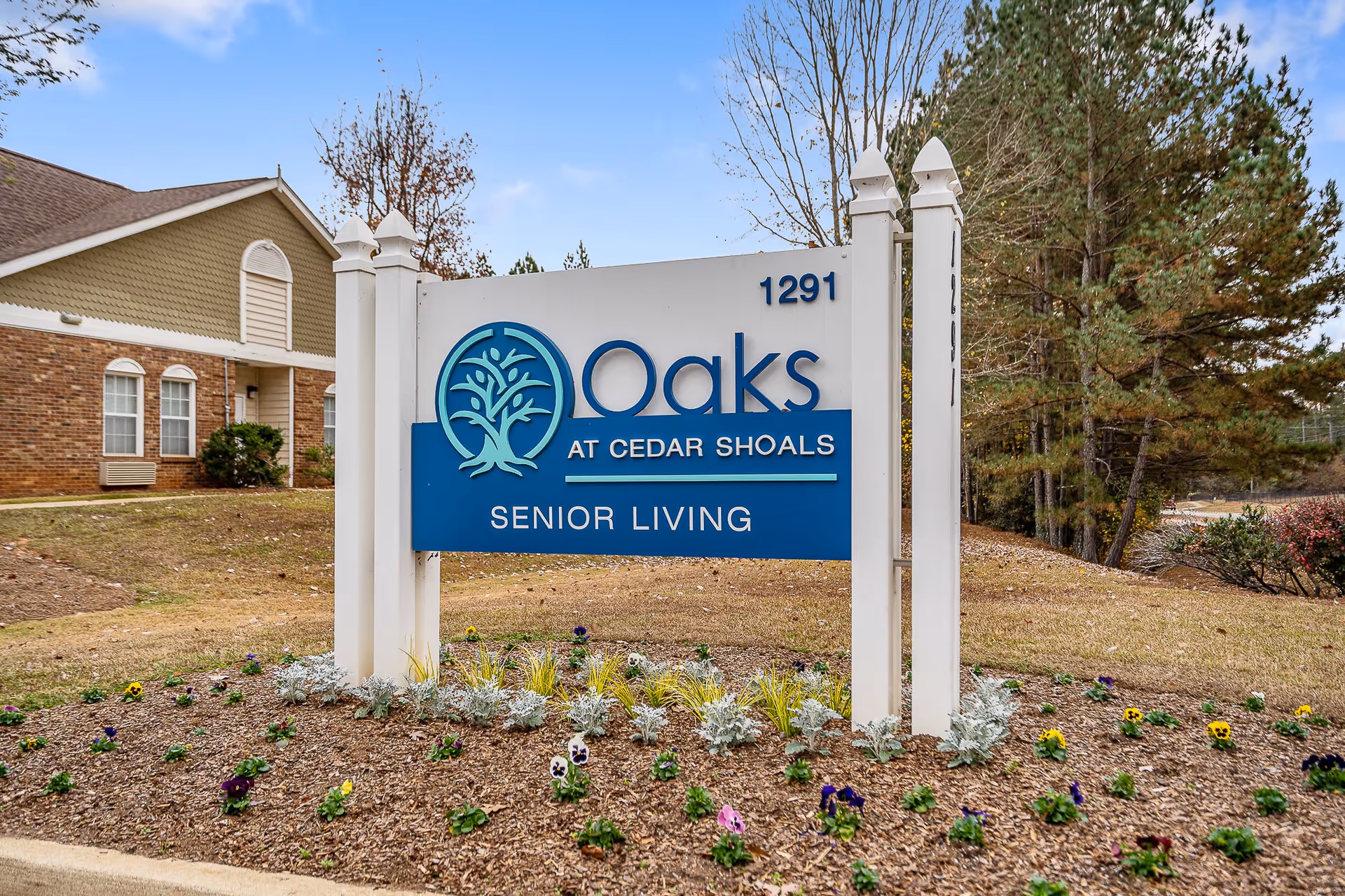 Outdoor view of a sign for Oaks at Cedar Shoals Senior Living, positioned on a landscaped area with flowers and mulch. In the background, there is a brick building and trees under a partly cloudy sky.